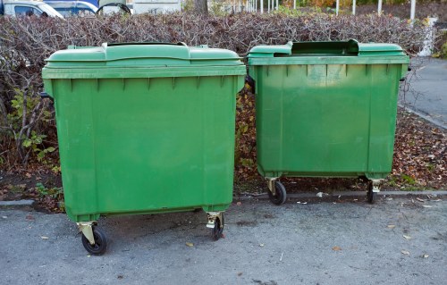 Two large green plastic wheelie bins with black wheels and closed lids are positioned on a paved driveway, side by side. The bins appear to be standard outdoor rubbish containers, with a smooth, slightly shiny surface and horizontal ridges near the top for structural rigidity. They are situated outdoors, with a background of low shrubs and leafless trees or bushes, indicating a typical residential area possibly during autumn or winter. The scene is evenly lit with natural daylight, showing clear details of the bins' textures and colors. This image exemplifies typical waste management equipment used by houses in the Camberley area, aligning with the services offered by House Clearance Camberley for rubbish removal and house clearance in Surrey, UK.