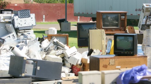 A large collection of discarded electronic waste, including old CRT computer monitors with wooden casings, boxed and unboxed computer components, and miscellaneous electronic devices, all piled on a paved outdoor area near a grassy verge with a brown fence and trees in the background. The electronic items vary in size, shape, and condition, with some stacked and leaning against each other, capturing the typical clutter associated with rubbish removal services in Camberley. Visible sunlight provides natural lighting, highlighting the mix of textures such as the smooth glass screens, worn plastic casings, and wooden finishes. The scene reflects a typical outdoor rubbish collection or clearance site, managed by companies like House Clearance Camberley, which specializes in removing household and electronic waste efficiently and responsibly in the local area.