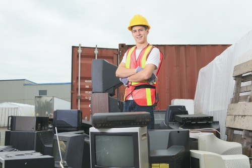 A young male worker wearing a yellow safety helmet and a high-visibility vest stands outdoors, smiling with arms crossed behind a collection of discarded electronic devices including old computer monitors, CPUs, and printers. The scene appears to be at a waste collection or disposal site, with a large red shipping container and a stack of wooden pallets visible in the background. The electronic waste is arranged in front of the worker on a paved surface, which suggests an organised rubbish removal operation. The overcast sky, combined with the industrial surroundings, highlights the focus on waste management services provided by House Clearance Camberley. This image illustrates a professional approach to rubbish collection, suitable for a site specialising in house clearance and electronic waste disposal, serving the local Camberley area.