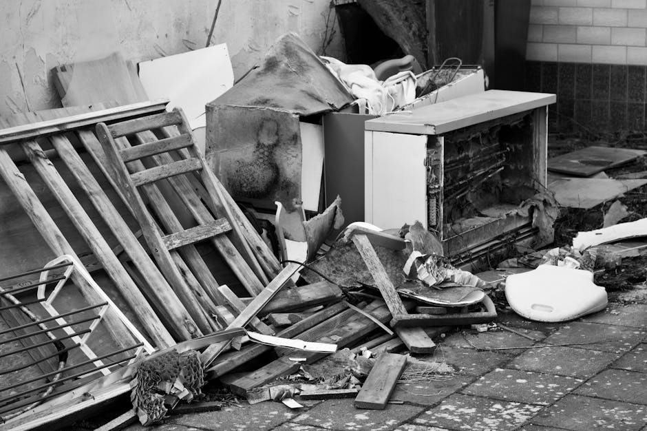 A large pile of discarded office furniture and equipment, including wooden desks, chairs, and metal-framed chairs, all scattered haphazardly on a warehouse or storage floor. The desks are light-colore