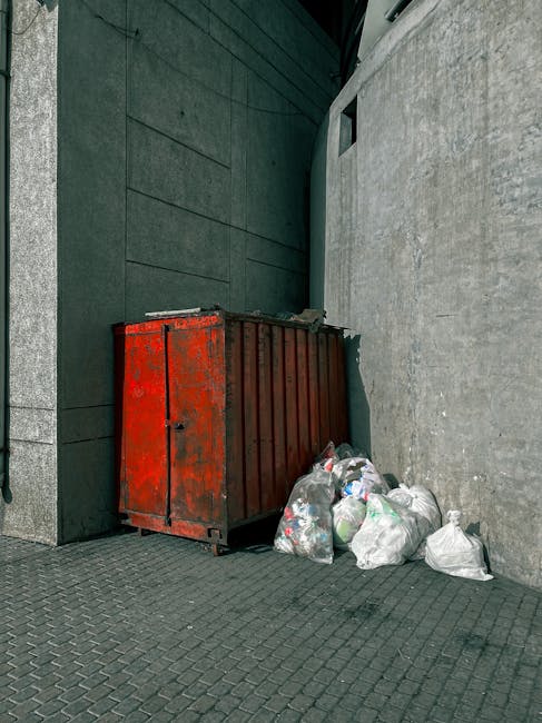 A large accumulation of black plastic rubbish bags stacked against a modern building with tiled exterior walls. The bags are filled with waste, and some are torn open revealing plastic bottles, paper,