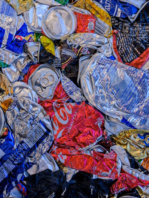 A close-up image of a large pile of crushed and crumpled aluminium beverage cans, predominantly silver in colour with some featuring branding and logos in red, blue, and black. The cans vary in size a